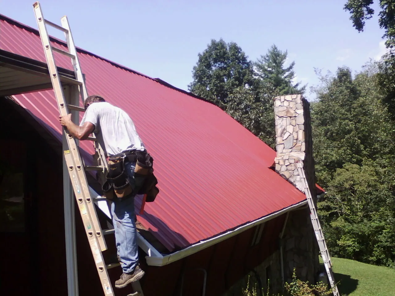 Roofer on a ladder beside a red metal roof and stone chimney — Soffit & Fascia Repair by Roofing Brothers in Clarkston